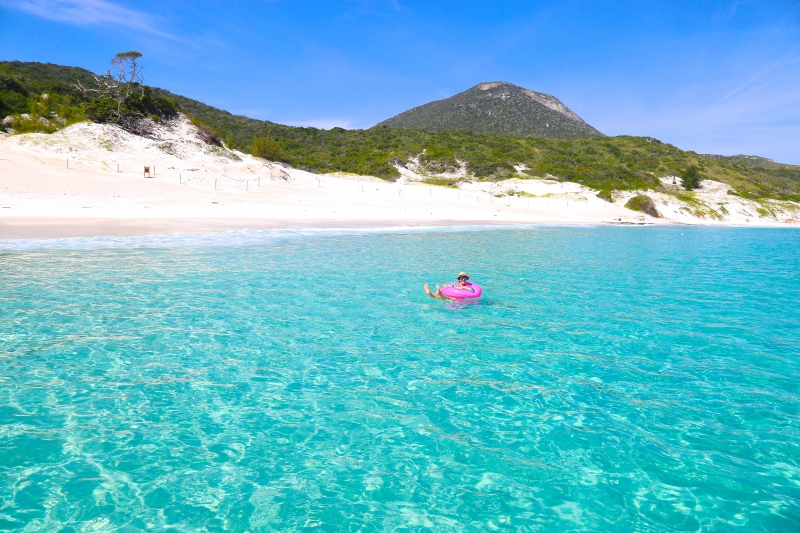 uma pessoa flutuando em um corpo de água com morros de areia e vegetação ao fundo 